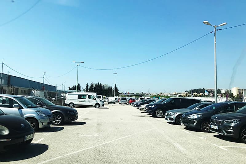 Offener Parkplatz am Hafen mit zwei Reihen geparkter Autos, einem weißen Lieferwagen und Laternen bei klarem Himmel.