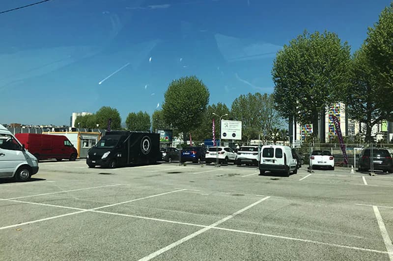 Freier Parkbereich mit weißen Parkmarkierungen, mehreren geparkten Fahrzeugen, Bäumen und Umzäunung im Hafen von Nizza.
