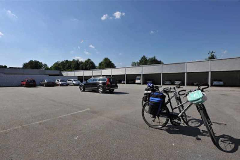 Große Freifläche eines Parkplatzes mit einigen Autos, überdachten Stellplätzen im Hintergrund und zwei Fahrrädern im Vordergrund.