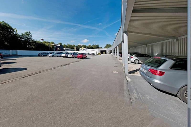 Großer Außenparkplatz mit überdachten Stellplätzen rechts, mehreren geparkten Autos und eingezäunter Umfriedung unter blauem Himmel.