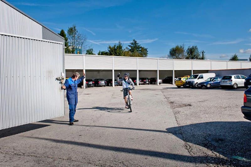 Parkplatz mit überdachten Stellplätzen, geparkten Autos, Fahrradfahrer und Mann in blauer Arbeitskleidung.