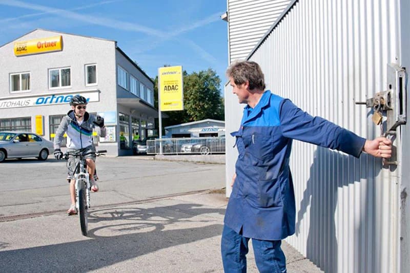 Mann in Arbeitskittel öffnet Tor, Radfahrer fährt auf dem Hof an, Autohaus mit ADAC-Schild im Hintergrund.