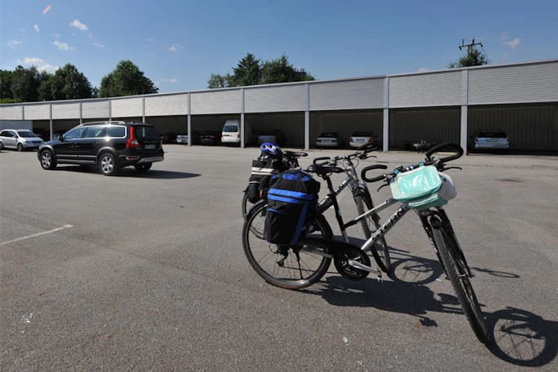 Großer Parkplatz mit überdachten Parkbuchten, mehreren Autos im Hintergrund und zwei beladenen Fahrrädern im Vordergrund.