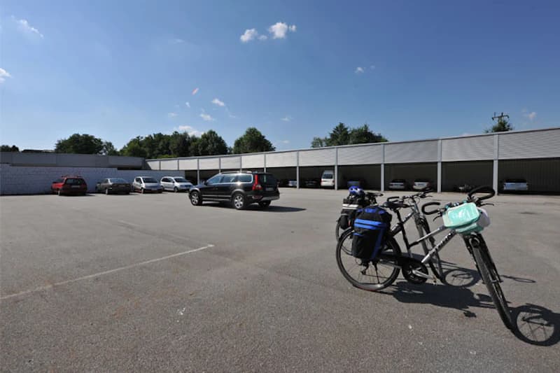 Großer Parkplatz mit zwei beladenen Fahrrädern im Vordergrund und überdachten Stellplätzen mit Autos im Hintergrund.