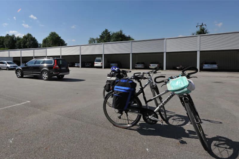 Zwei Fahrräder mit Taschen auf einem großen Parkplatz vor überdachten Stellplätzen und geparkten Autos.
