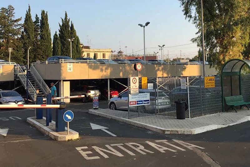 Zweistöckiges Parkhaus mit offener Zufahrt, Schranke, Umzäunung, Lampen und geparkten Autos.