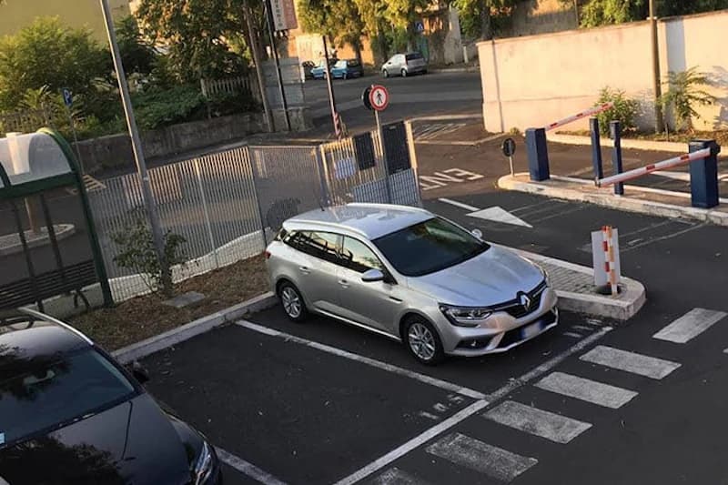 Silbernes Kombi in markiertem Parkplatz neben Gitterzaun und Schranke am Einfahrtsbereich, Zebrastreifen sichtbar.