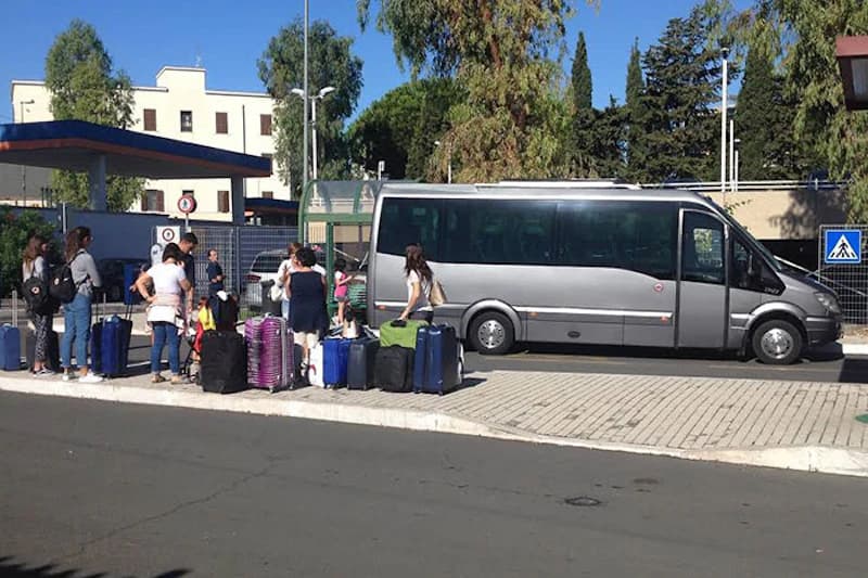 Gruppe von Reisenden mit Koffern wartet am Gehsteig neben einem silbernen Kleinbus vor Zaun und Bäumen.