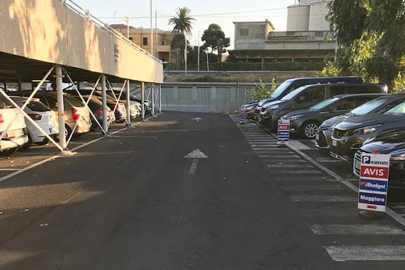 Parkplatz mit geparkten Autos, überdachte Stellplätze links, reservierte Parkschilder rechts und Richtungspfeil auf dem Asphalt.