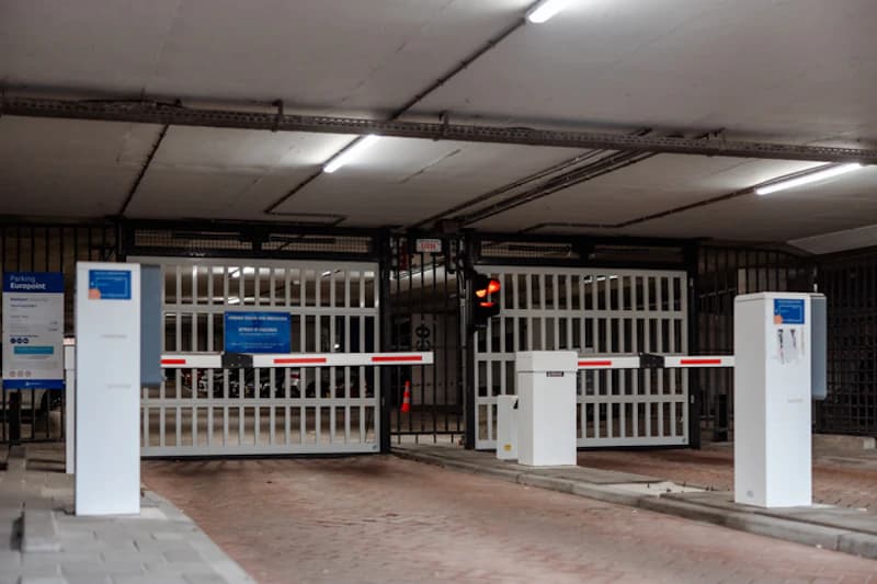 Überdachte Parkhauseinfahrt mit Schranken, geschlossenen Metalltoren, Kassenautomaten und roter Ampel.