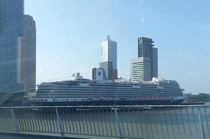 Großes Kreuzfahrtschiff am Kai im Hafen von Rotterdam mit Hochhäusern im Hintergrund und Geländer im Vordergrund.