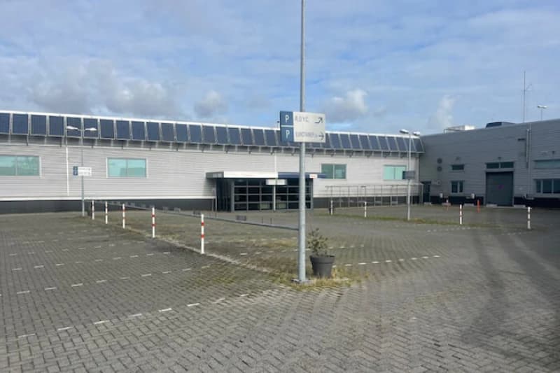 Leerer gepflasterter Parkplatz mit Markierungen, Pollern, Laterne und Industriegebäude mit Solarzellen.