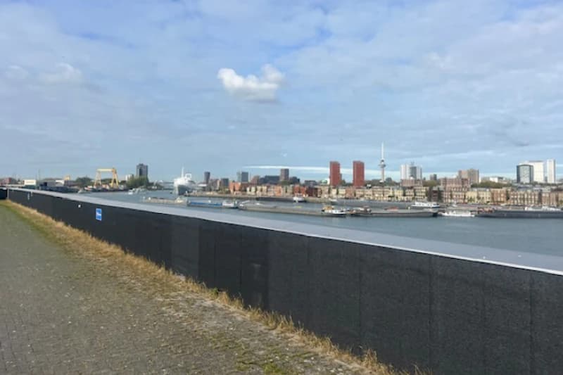 Promenade und Schutzmauer am Wasser mit Schiffen und der Skyline von Rotterdam im Hintergrund.