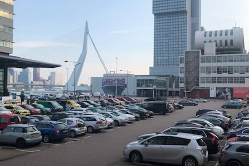 Großer Parkplatz am Hafen mit dicht geparkten Autos und der Erasmusbrücke im Hintergrund.