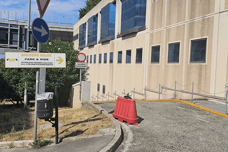 Zufahrt mit rotem Kunststoff-Absperrblock, Verkehrsschildern, Hinweisschild, Papierkorb und heller Gebäudefassade.