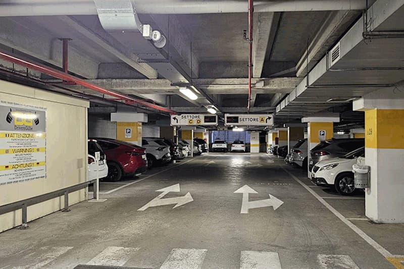 Parkhalle mit Fahrpfeilen auf dem Boden, geparkten Autos an beiden Seiten, gelbweißen Stützen und Deckenbeleuchtung.