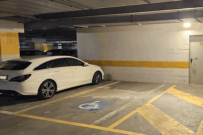 Überdachte Parkebene mit weißem Kombi und markiertem Behindertenparkplatz neben Wand mit gelbem Streifen.