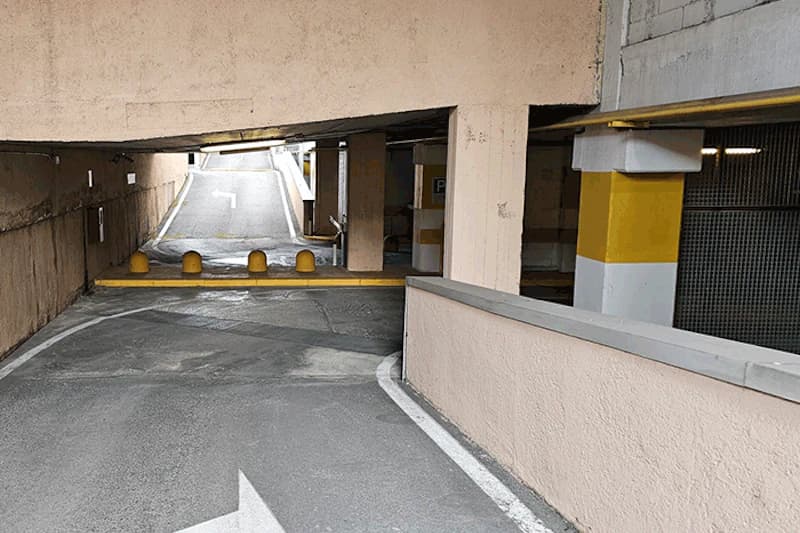 Abwärtsrampe einer Parkhalle mit gelber Schrankenbake, vier runden Pollern und Pfeilmarkierung auf dem Asphalt.