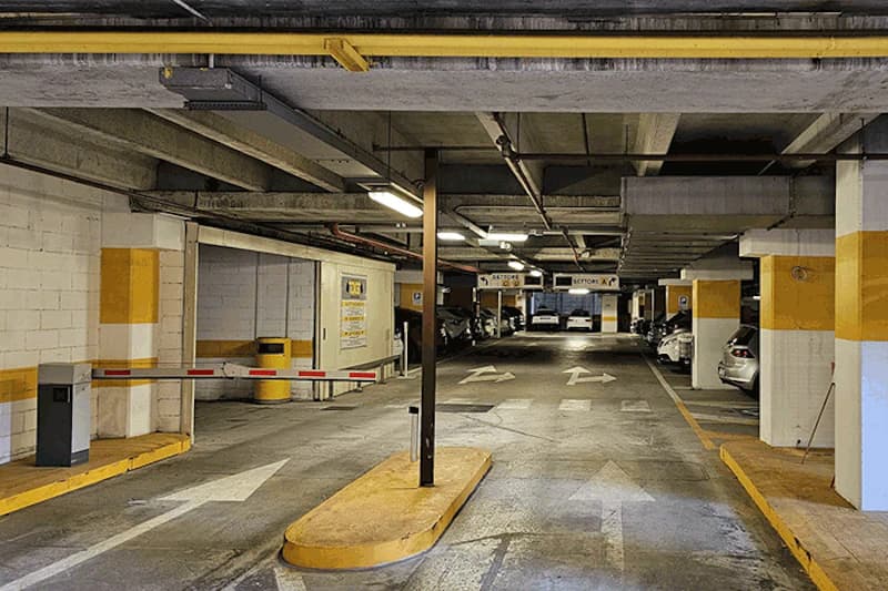 Überdachte Parkhalle mit Schranke, Fahrbahnmarkierungen und geparkten Autos zwischen gelb markierten Stützen.