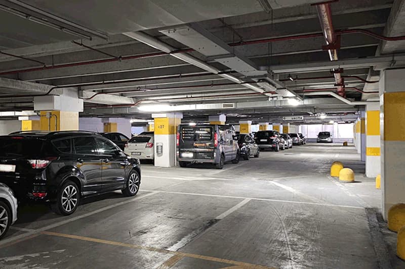 Blick in eine beleuchtete Tiefgaragenhalle mit parkenden Autos, gelb markierten Stützen und Deckenleitungen.