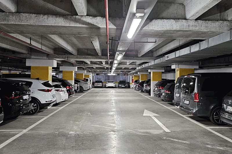 Tiefgaragenhalle mit geparkten Autos beidseitig, Betonstützen mit gelben Markierungen und Deckenbeleuchtung.