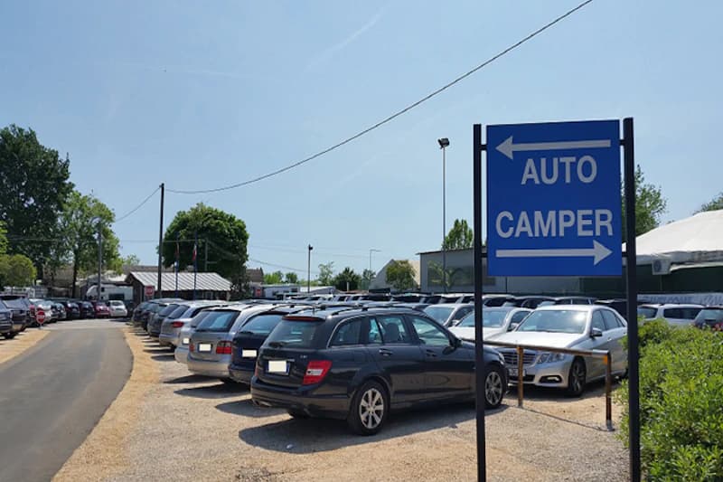 Kiesparkplatz mit dicht geparkten Autos, blaues Schild mit der Aufschrift AUTO CAMPER und Richtungspfeilen.