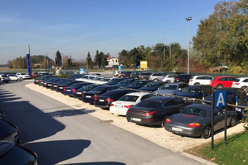 Außenparkplatz mit dicht geparkten Pkw in Reihen, Bereichszeichen A, Beleuchtungsmasten und Bäume im Hintergrund.