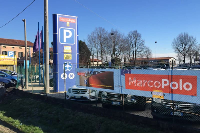 Eingezäunter Parkplatz mit hoher blauer Parktafel, MarcoPolo-Banner und geparkten Autos hinter Drahtzaun.