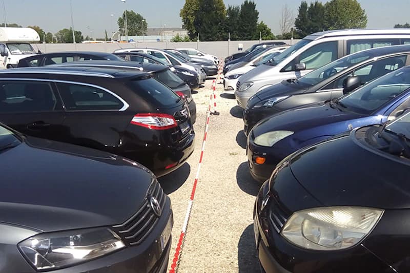 Zwei Reihen geparkter Autos auf einem Schotterparkplatz, in der Mitte rotweiße Absperrkette, Zaun und Bäume im Hintergrund.